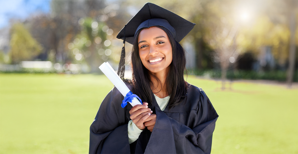Smiling graduate in cap and gown holding a diploma tied with a blue ribbon, standing outdoors on a sunny day.