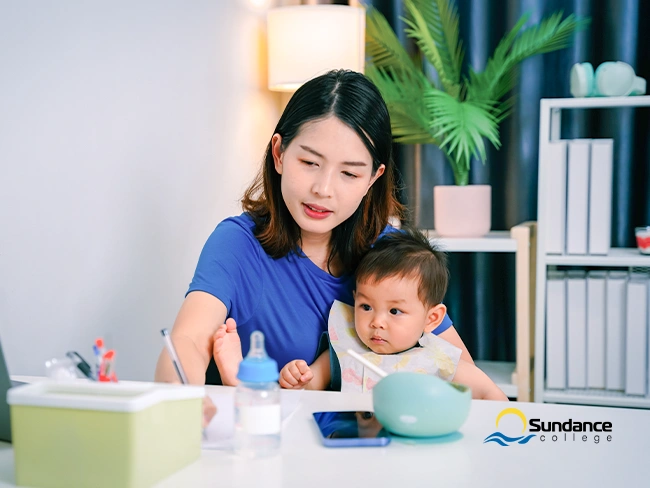 Mother studying at home with her baby on her lap, multitasking with a laptop, bottle, and study materials on the table.