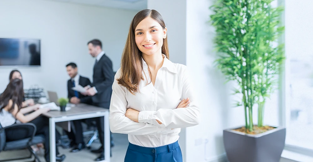 Smiling HR coordinator standing confidently in a modern office, while a business team collaborates during a meeting in the background.