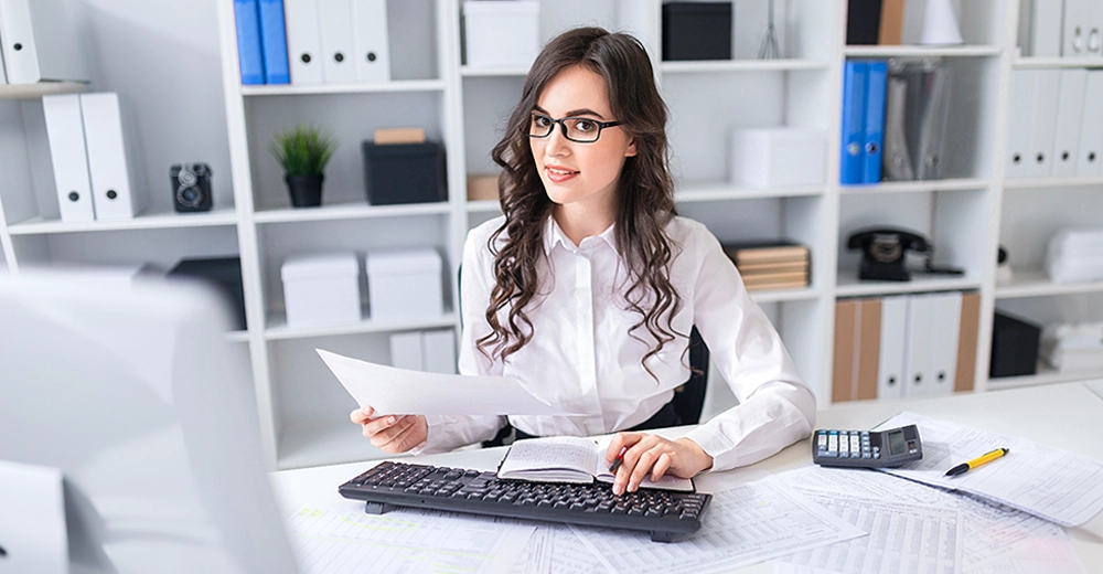 Woman sitting behind a computer desk glancing to the right with paperwork and a calculator.
