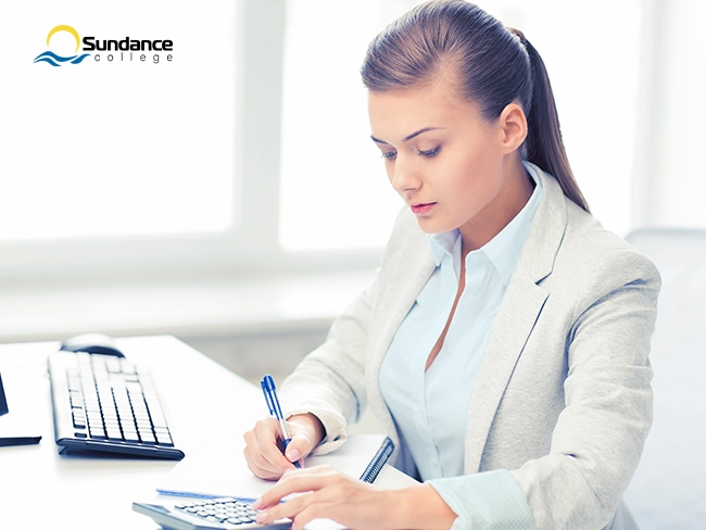Woman sitting behind a computer desk using a calculator while writing details into notebook.
