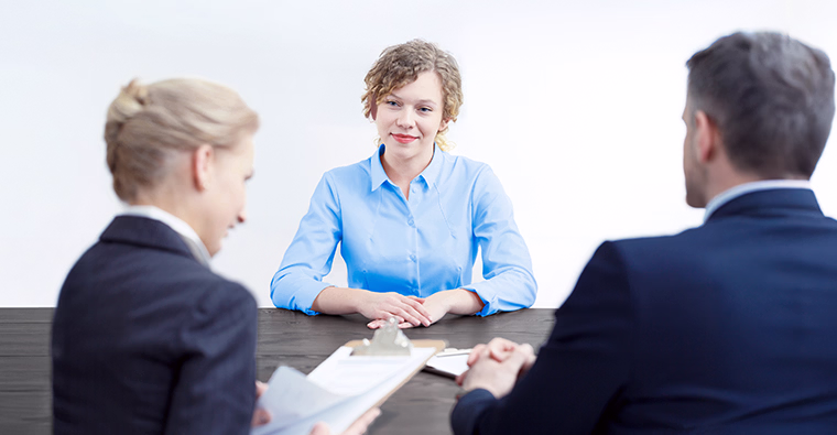 A woman sits at a table, with two interviewers who are asking HR management interview questions.