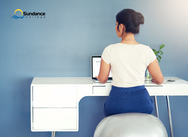 Professional woman sitting on a grey exercise ball at her desk, typing on a computer with her back facing the camera.
