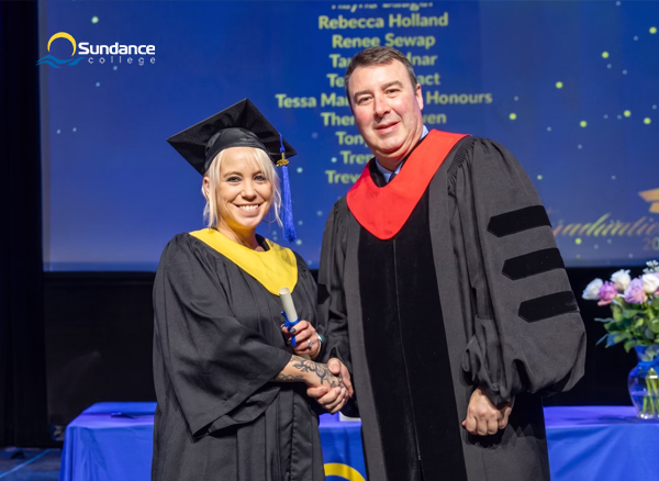 Shelley B., an Addictions and Community Health Professional Diploma graduate, standing on the stage with Dean Olsen, President of Sundance College, during the graduation ceremony in Winnipeg.