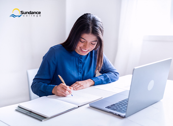 A young woman in a sweater sits smiling in front of a laptop as she writes in a notebook.