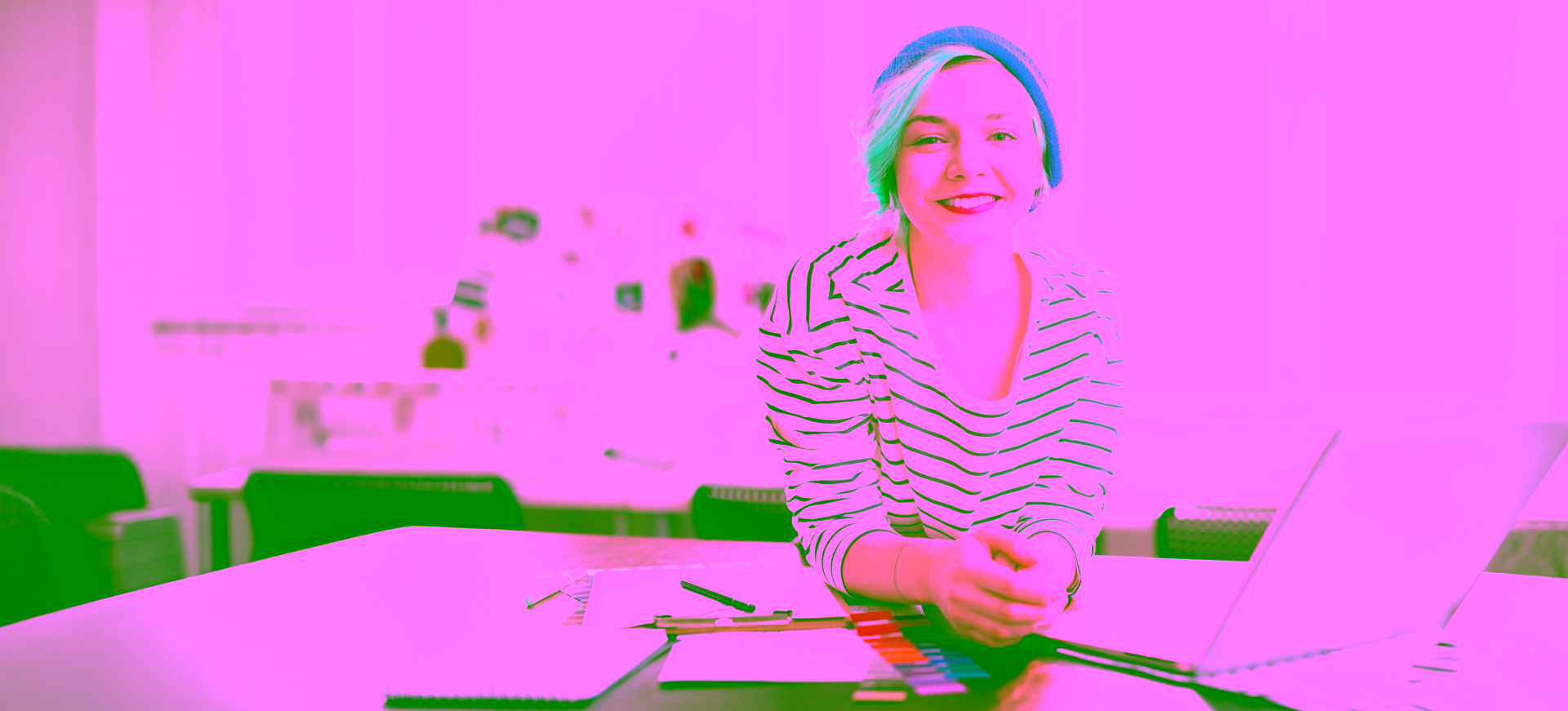 A female creative designer at a studio leans over her wooden work desk, directly facing the camera with her hands clasped. The work desk is full of design colour swatches, notebooks, and a laptop.
