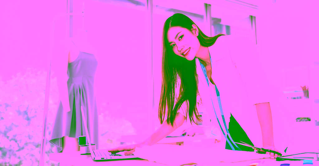 A smiling fashion designer leans on her workbench, a tape measure draped over her shoulders.