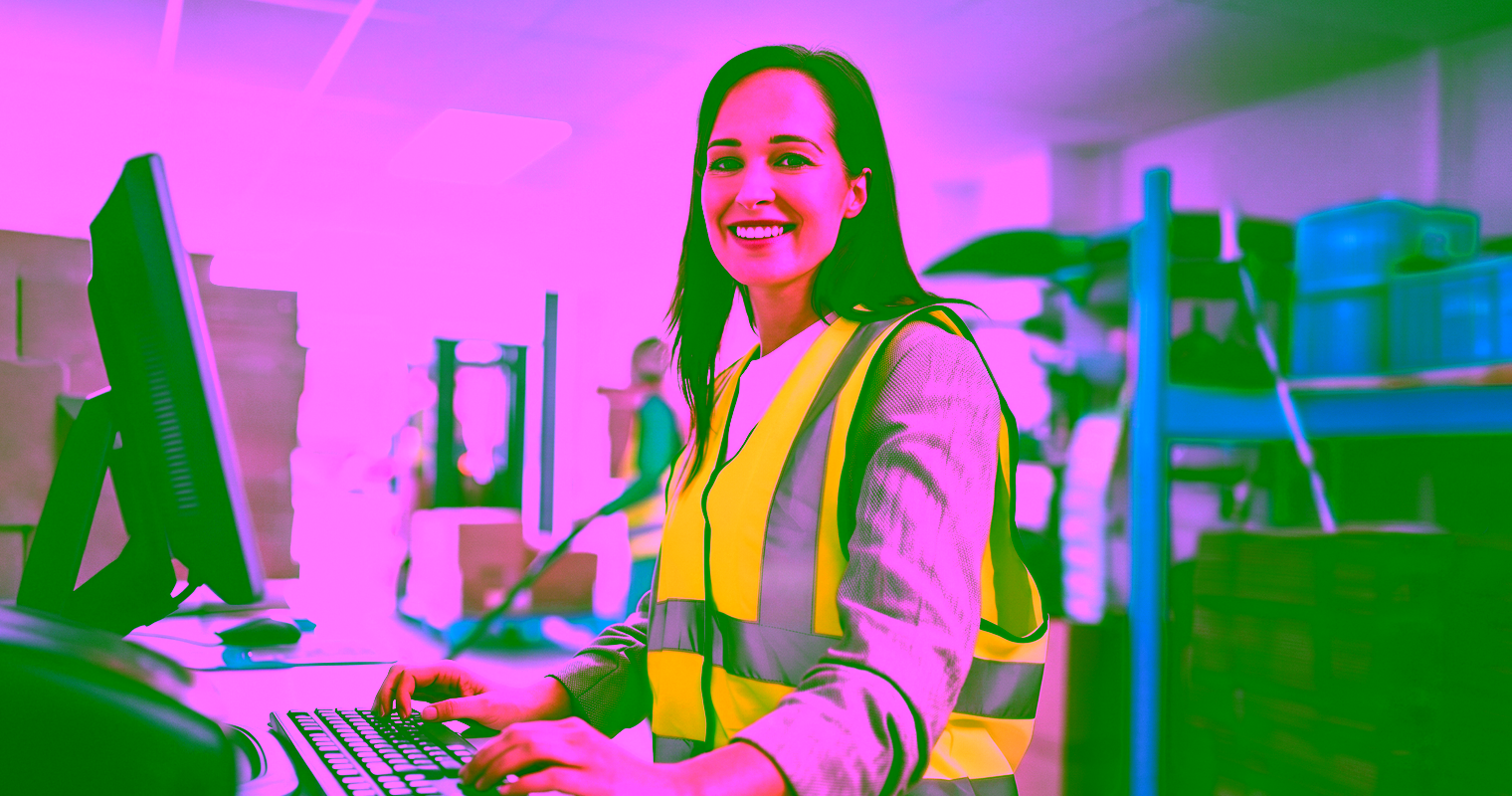 A female supply chain worker wearing a high-visibility safety vest typing at a computer terminal in a warehouse.