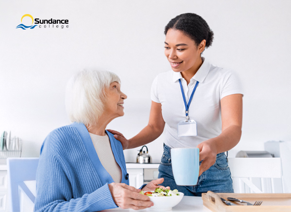 Personal support care worker in a dining room giving an elderly woman a mug of tea as she eats a salad for lunch.