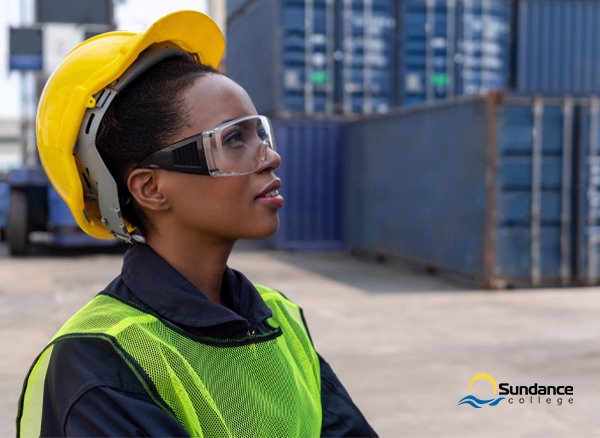 A female supply chain and logistics professional wearing a yellow hard hat and safety glasses in front of shipping containers.